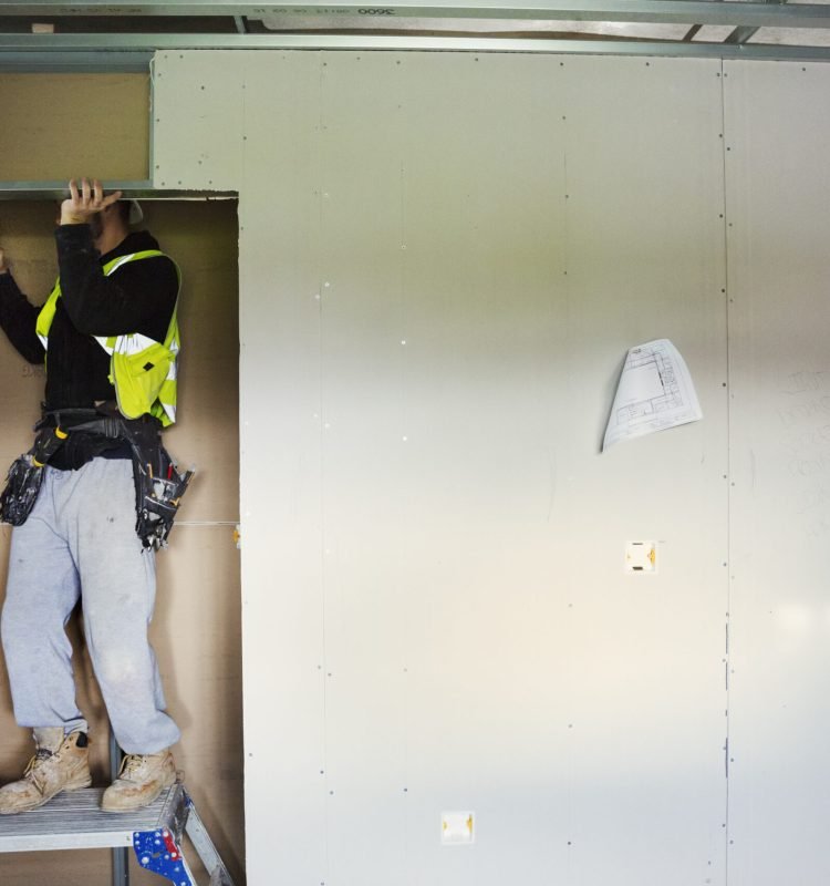 A carpenter in work clothes on a construction site, using an electric drill.