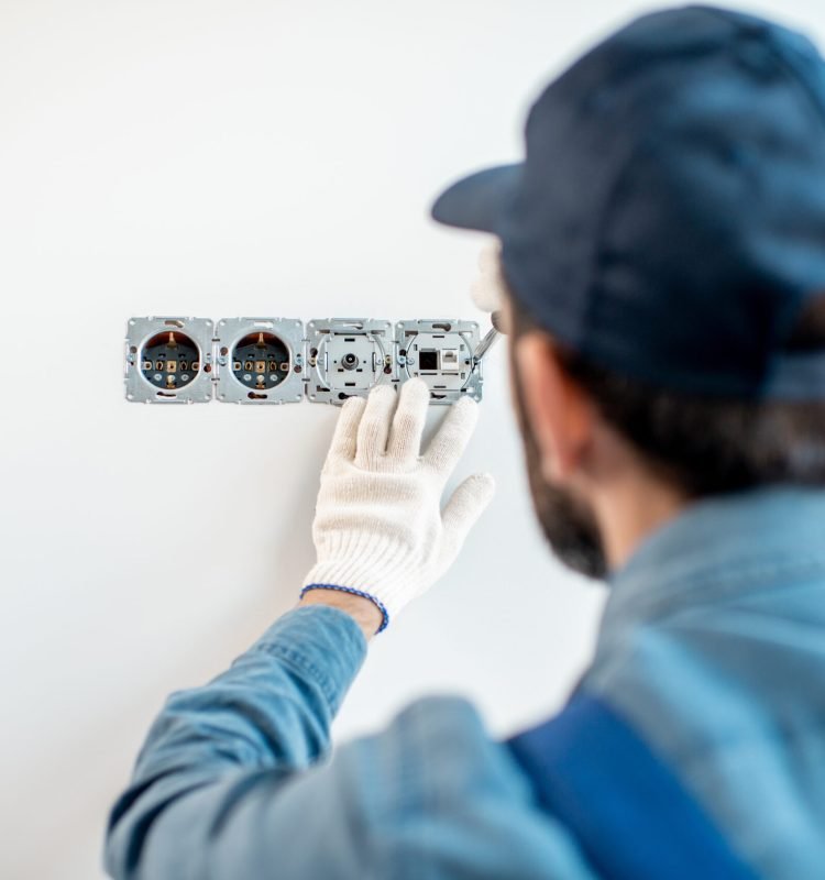 Electrician in uniform mounting electric sockets on the white wall indoors