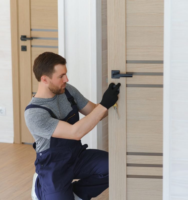 Installation of a lock on the front wooden entrance door. Portrait of young locksmith workman in blue uniform installing door knob. Professional repair service. Maintenance Concept.