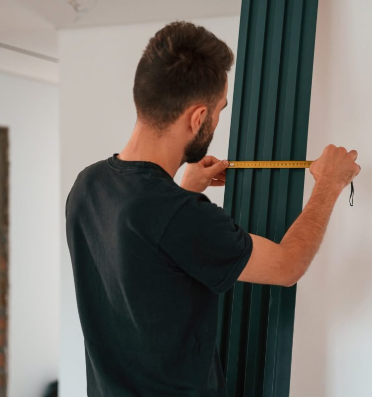 Taking measurements. Plumber worker installing heating radiator in empty room of a newly built house.