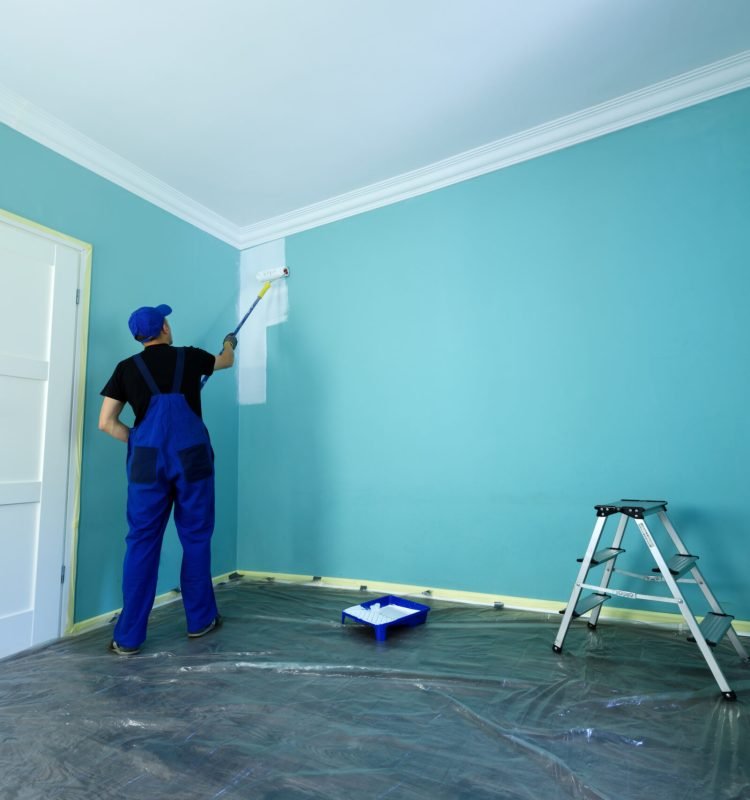 Man in a working overall is painting the wall in white color
