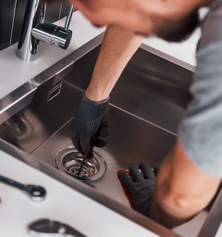 Young professional plumber in grey uniform working on the kitchen.