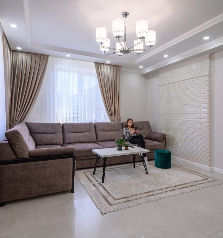 Young cute female using her smartphone while sitting at sofa in front of window in studio apartment interior
